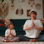 A mother and her small daughter are bonding and practicing yoga in the living room at home.